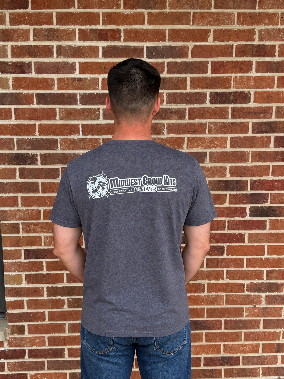 Person wearing a gray t-shirt with a logo and text, standing against a brick wall.