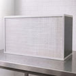 A rectangular white FFU with pleated air filter with a white frame, resting on a stainless steel workbench against a white brick wall background.