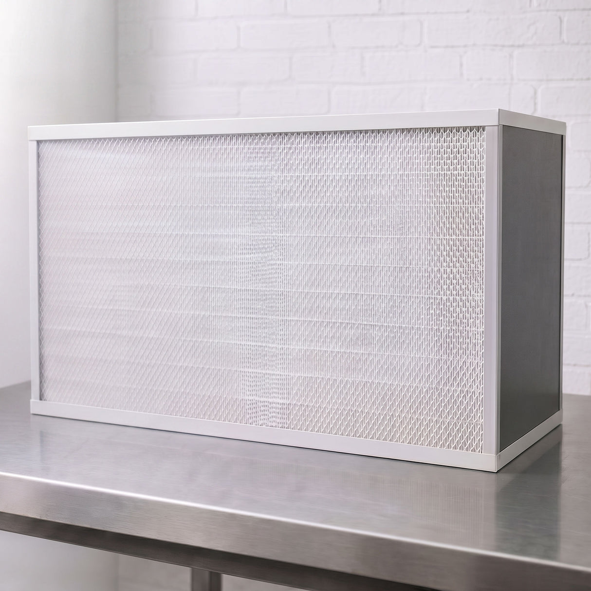 A rectangular white FFU with pleated air filter with a white frame, resting on a stainless steel workbench against a white brick wall background.