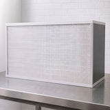 A rectangular white FFU with pleated air filter with a white frame, resting on a stainless steel workbench against a white brick wall background.