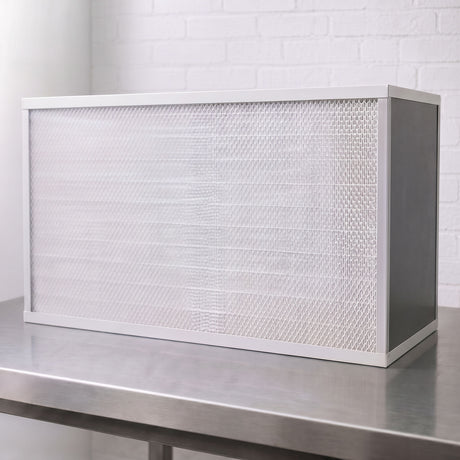 A rectangular white FFU with pleated air filter with a white frame, resting on a stainless steel workbench against a white brick wall background.