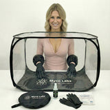 A woman wearing a pink top and black gloves stands behind a clear pop-up containment tent with black trim. In front of her are additional included items: a black zippered pouch, a small spray bottle, a pair of black gloves.