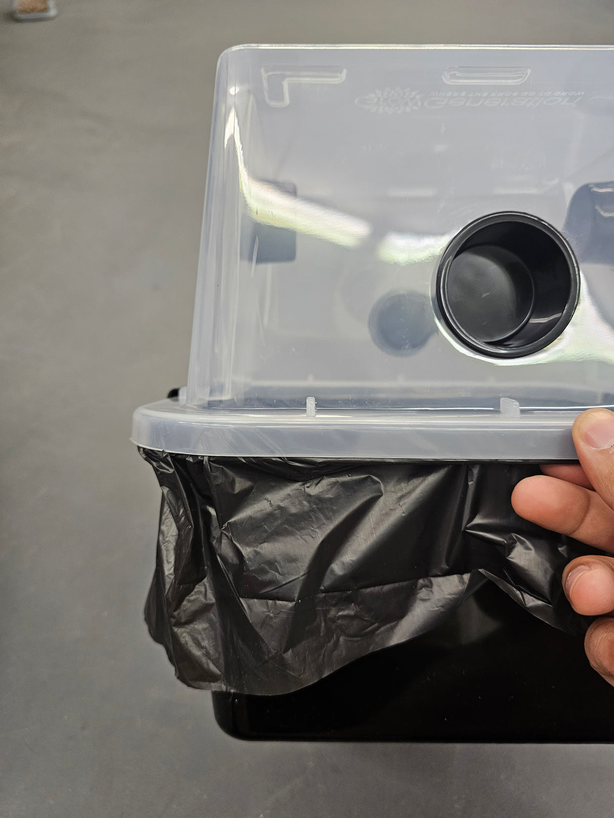 A hand is holding a clear plastic storage container with a black circular opening, showing a black plastic liner or bag being fitted inside.
