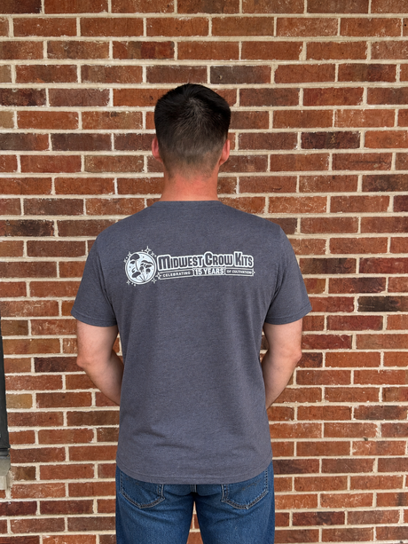 Person wearing a gray t-shirt with a logo and text, standing against a brick wall.